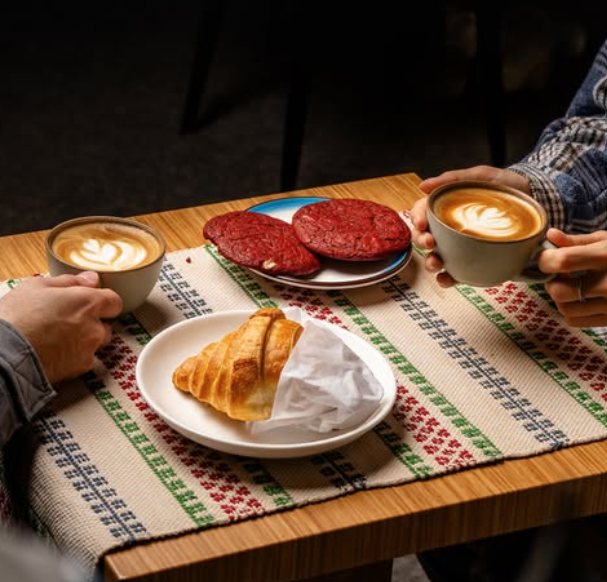 Two lattes with red velvet cookies and a croissant on a woven table runner